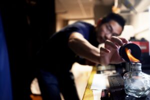 A man leans over a lab bench as he positions a thin, transparent film into a donut-shaped clamp. The camp is positioned so that the hole faces a flame dancing atop a small oil burner.