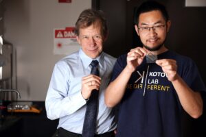 A man in a light-blue shirt and dark blue tie stands next to another man in a dark blue graphic T-shirt. Both are holding a thin, transparent film cut into a small square.