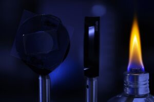 Two donut shaped clamps hold a thin, transparent film inside a dimly lit lab. One donut is pointed toward a tiny, orange flame burning on a small, candle-like object.
