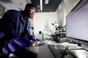 A scientist stands at a lab bench and reviews data on a computer monitor. A black platform containing the heat source and photovoltaic cell sits next to the monitor.