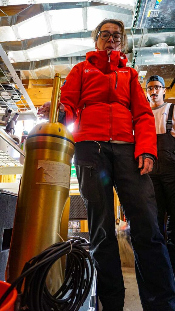A woman stands with one hand on a cloud imaging probe, which looks like a golden cylinder that stands to about hip height. The probe has a smaller, antenna-like cylinder that extends from the domed top of the probe.