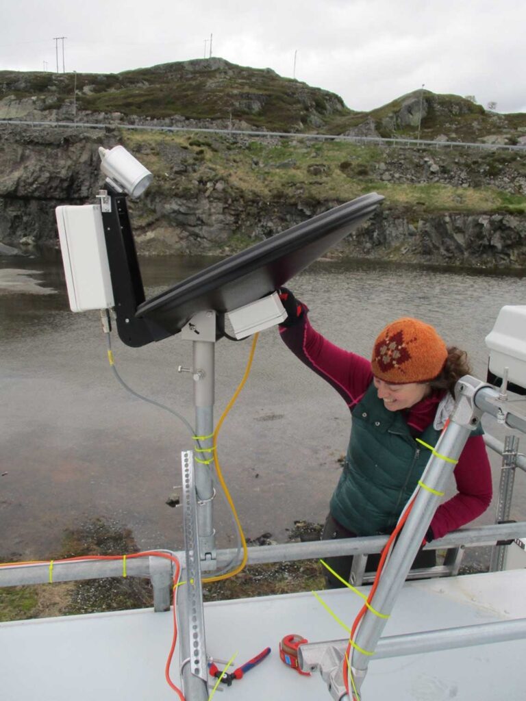 A black radar dish is mounted on a metal pole on the roof of a building. A scientist in winter clothes has a hand on the dish and stands on a ladder leading down from the roof.