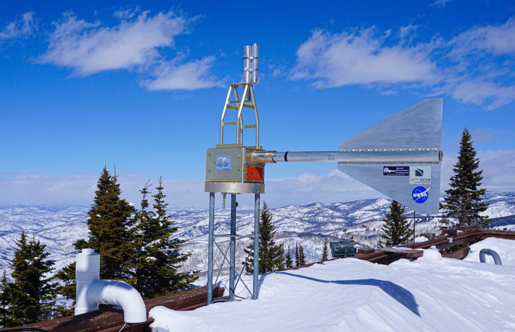 A finned cylinder protrudes from a metal box situated on a tower on the snow-capped roof of a laboratory. The lab and tower overlook a mountainous landscape covered in bright white snow.