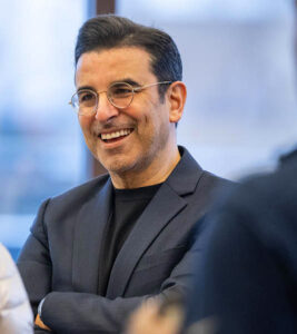 A man with glasses smiles in mid-conversation.