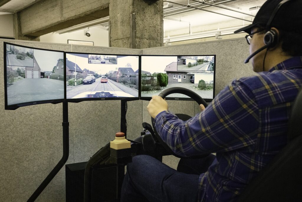 A man sits in front of a bank of three computer screens, holding a steering wheel as he pilots a remote vehicle.