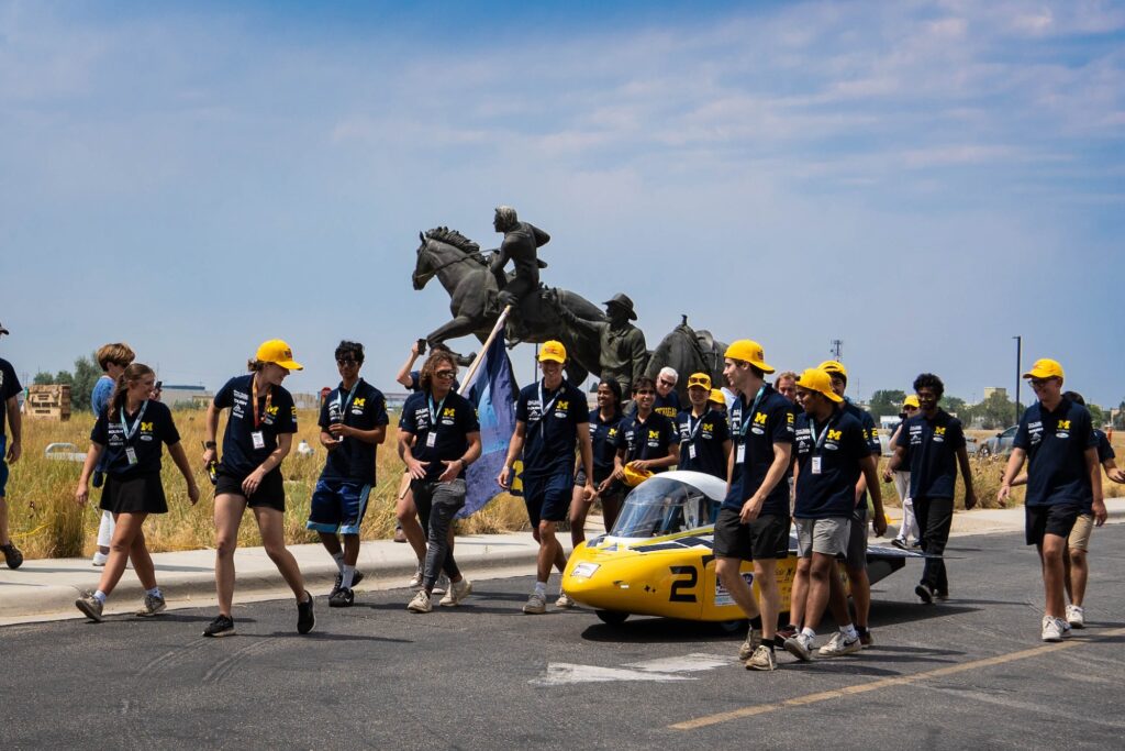 College students surround a custom vehicle they built as it crosses a race finish line.