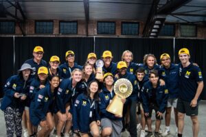 College students smile with their arms around each other. One holds a gold trophy.