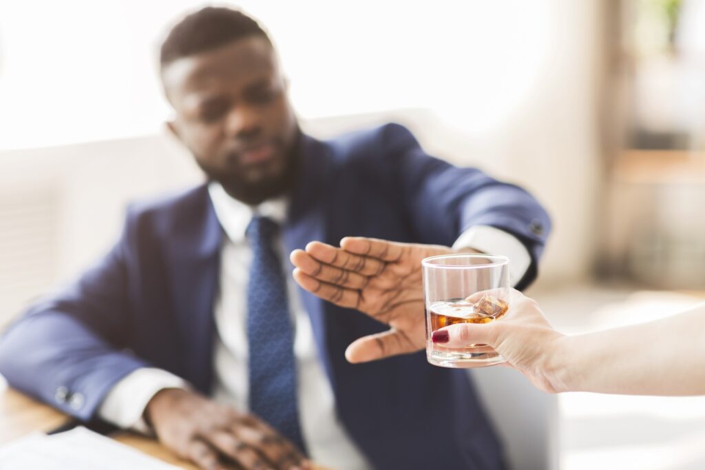 African American businessman saying no to glass of whiskey offered by female colleague, selective focus, copy space. Image credit: iStock