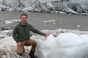 As Mark kneels, the block of ice comes to about waist height. The ice sits on a rock near the coast, and its rounded edges suggest it is melting.