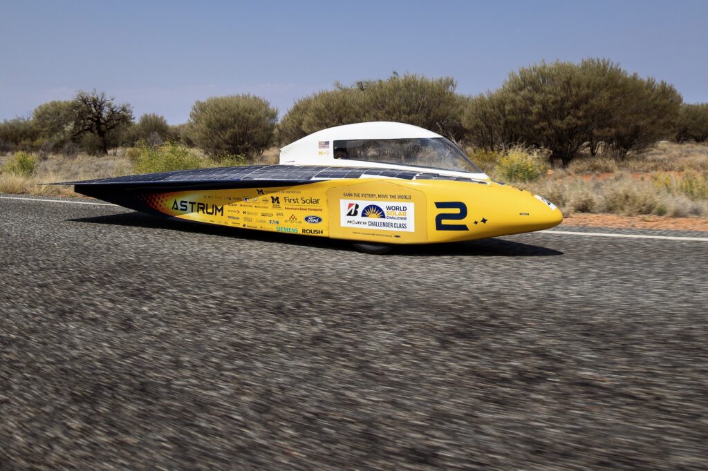 A car shaped like a yellow bullet with a white cockpit and solar panels on top moves along a black-paved, Australian road.