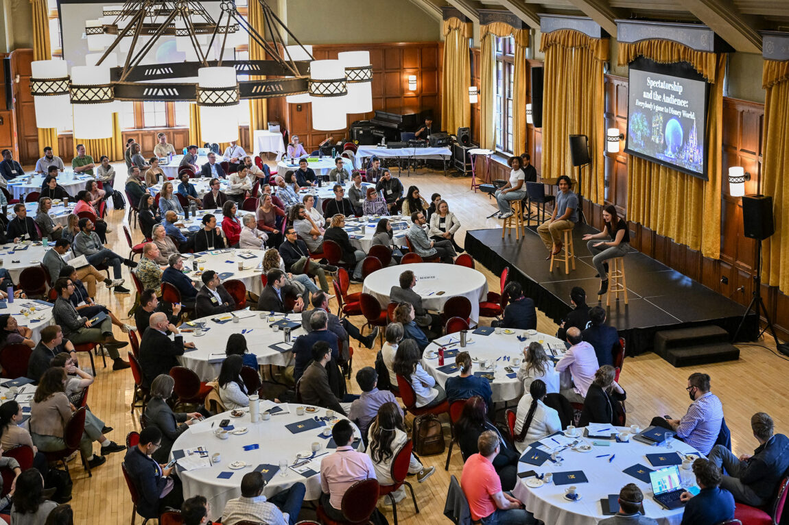 Overview shot of people sitting at tables watching three people on a stage