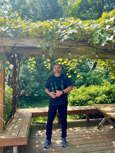 A photo of a man standing in an orchard holding bottles of homemade wine