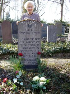 A white-haired woman, Irene Hasenberg Butter, stands behind the tombstone of her father, which is printed with Hebrew writing. She is in Laupheim, Gemany.