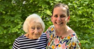 Two women, one who is elderly, stand together in front of greenery. The elderly woman was a pioneering female professor at U-M School of Public Health; the younger woman is a currently faculty member. Their families are connected because of the Holocaust.