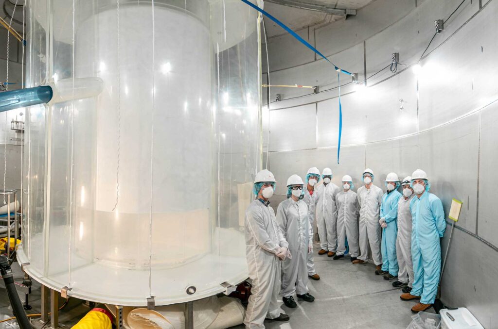 A group of nine researchers wearing white or blue clean room garments with facemasks, hairnets and hard hats stand in a white room near a large cylindrical instrument used in the search for dark matter.