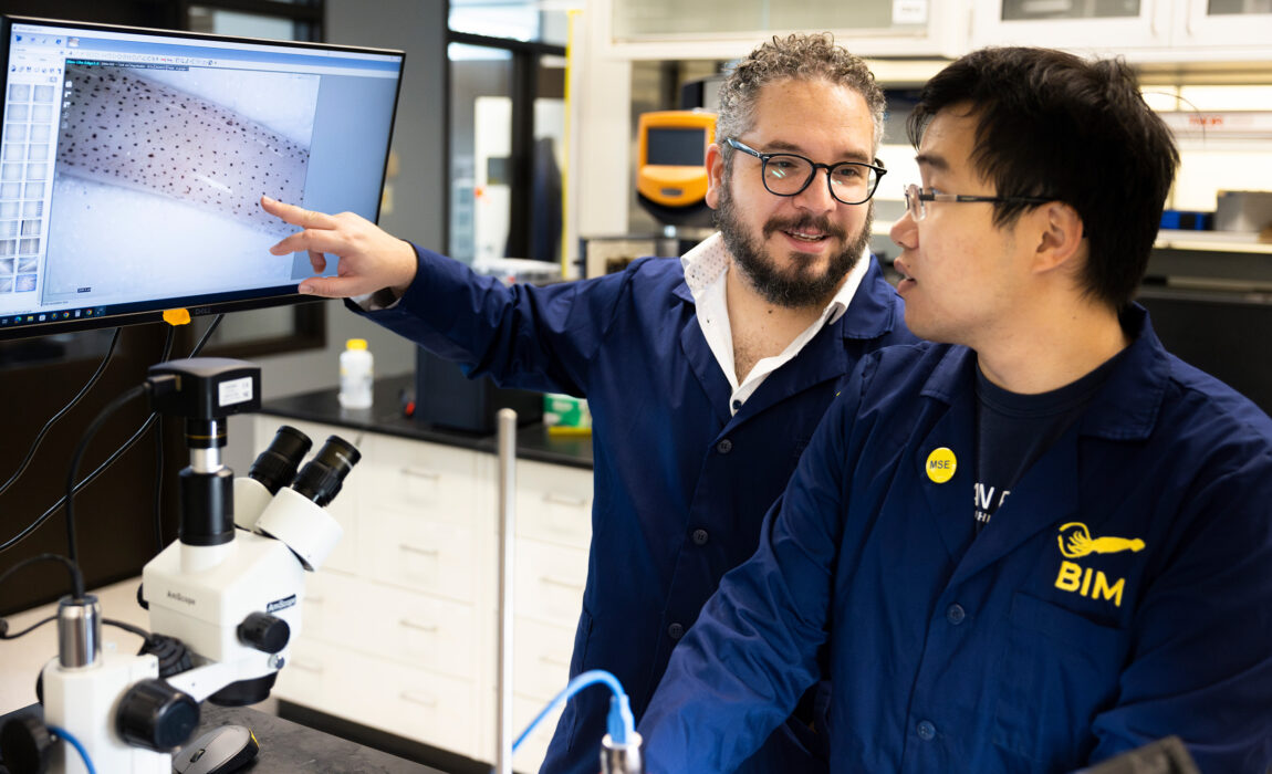 One man points at a screen while talking to another in a lab.