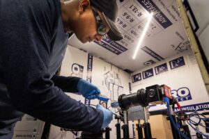 A researcher wearing safety glasses, gloves and a baseball cap connects a wire to the OLED, two sheets of glass about ten by ten centimeters with translucent gray strips sandwiched between. A mounted, horizontally positioned microscope points at the OLED. Housing insulation surrounds the experimentation area.