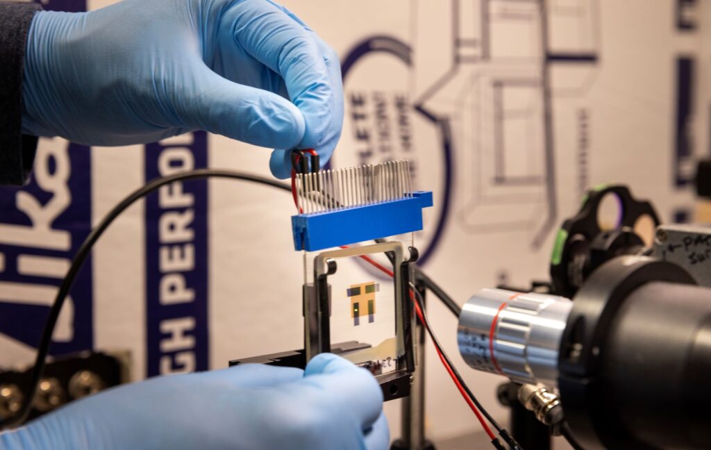 Gloved hands connect a wire to the OLED, two sheets of glass about ten by ten centimeters, with four, one centimeter long translucent gray strips sandwiched between. The silver barrel of a microscope points at the OLED. Housing insulation is out of focus behind.