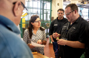 A woman speaks with three men—the whole group wearing safety glasses—discussing data on a sheet of paper at a battery training event.
