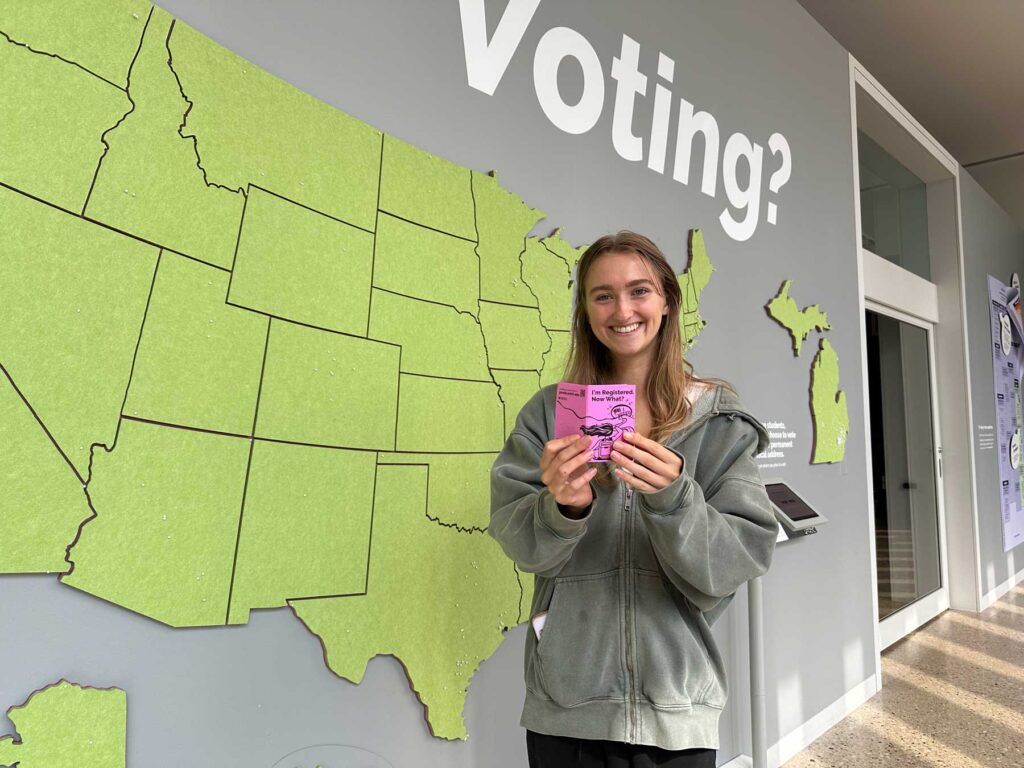 A college-aged woman faces the camera smiling,
Holding out a small pink folded paper Zine, entitled “I’m registered. Now what?” Behind her, a large green felt map of the United States and the State of Michigan hangs on the wall, stuck with small white pins in various locations. Large white text overhead reads, “Voting?”