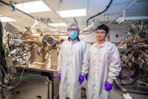 Pan and Bingxing stand in a lab. Two devices in the background are full of portholes, pipes and wires.