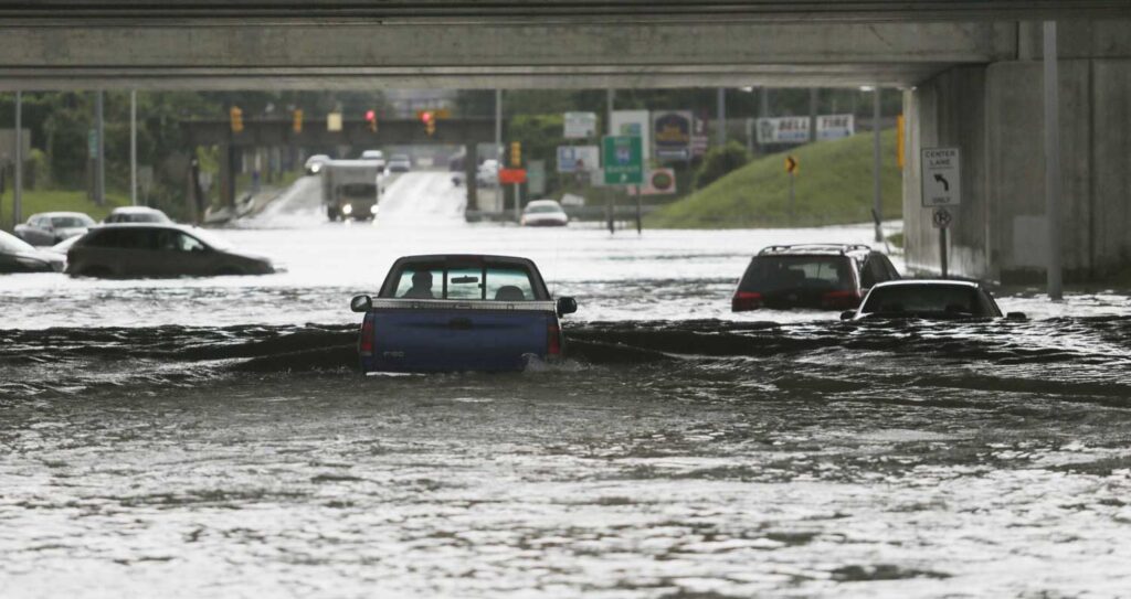 A pickup truck, submerged in water halfway up the driver’s door, is seen from the back attempting to drive beneath an overpass, while nearby cars appear stuck in the flooding.