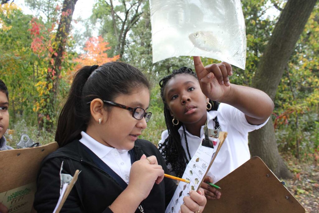 Two students discuss worksheets they're looking over while standing in a wooded area.