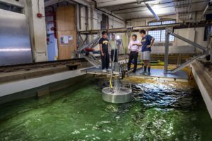 Four men stand on a platform stretching across a large indoor tank of water. A light shines on a metal buoy floating in the tank near the group.