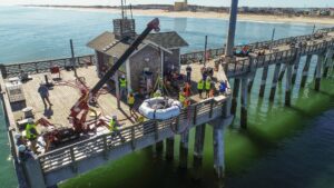 A crew of construction workers lower a wave energy device into the ocean. The device resembles an inner tube with machinery in the center seat.