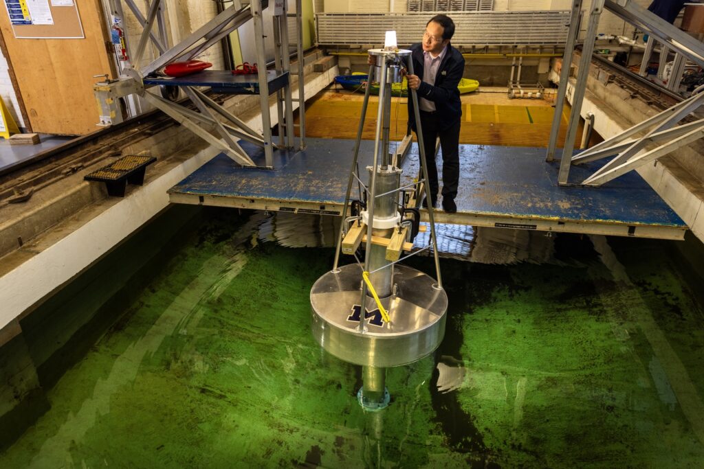 A metal buoy floats inside an indoor tank of water. A man closely inspects an illuminated light bulb attached to the top of a post on the buoy.