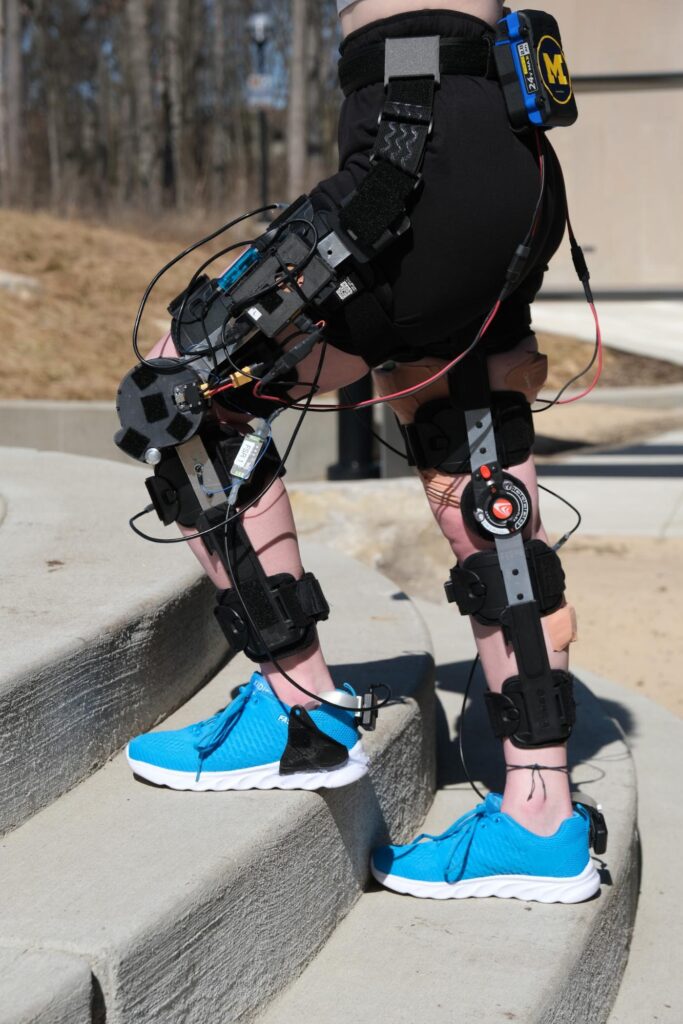 Knee braces wrap around each leg, from the upper thigh to lower calf. A flat, round motor is positioned on the outside of the knee where the brace hinges. Wires run up and down the legs, some running to a battery positioned on the participant's lower back.