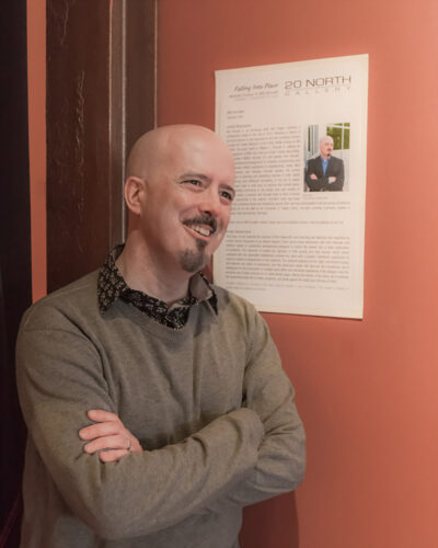 A photo of a man leaning against a wall with his arms crossed.
