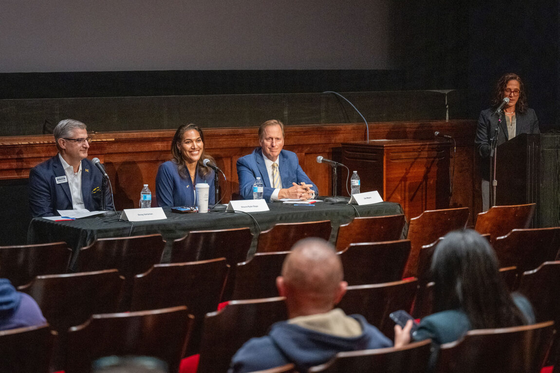 Photo of two men and a woman sitting at a table with another woman standing at a microphone