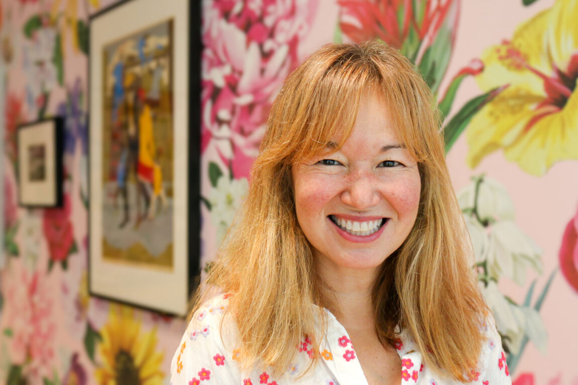 A photo of a smiling woman in front of a large floral-print wall and framed photos.