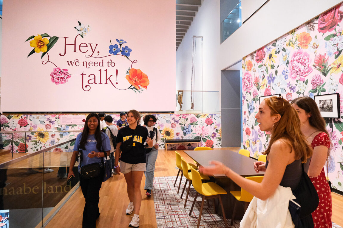 Photo of students walking through a gallery exhibit with large floral-print walls and a sign that reads, "Hey, we need to talk!"