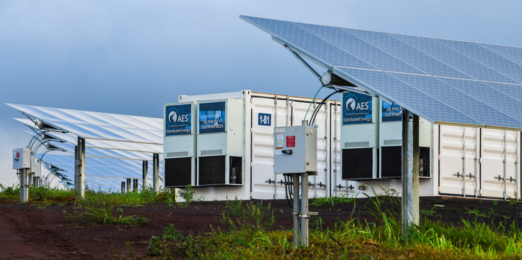 Two shipping-container sized battery energy storage systems sit between arrays of solar panels.