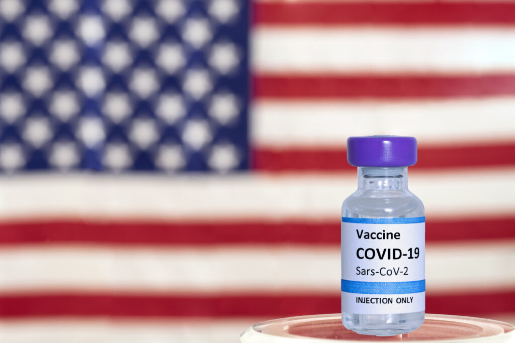 Vial of COVID-19 vaccine with an out of focus American flag in the background. Image credit: iStock