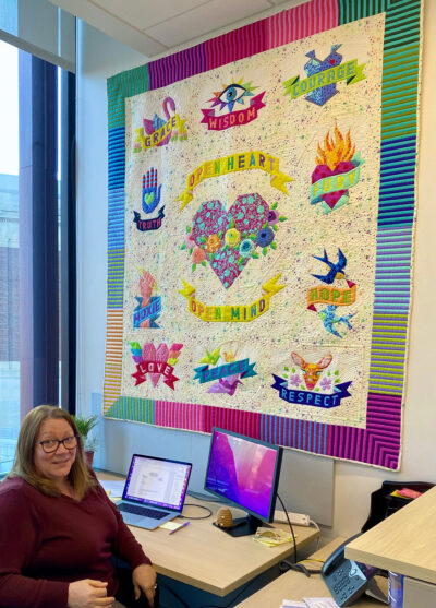 A photo of a woman at her desk, above which hangs a large quilt