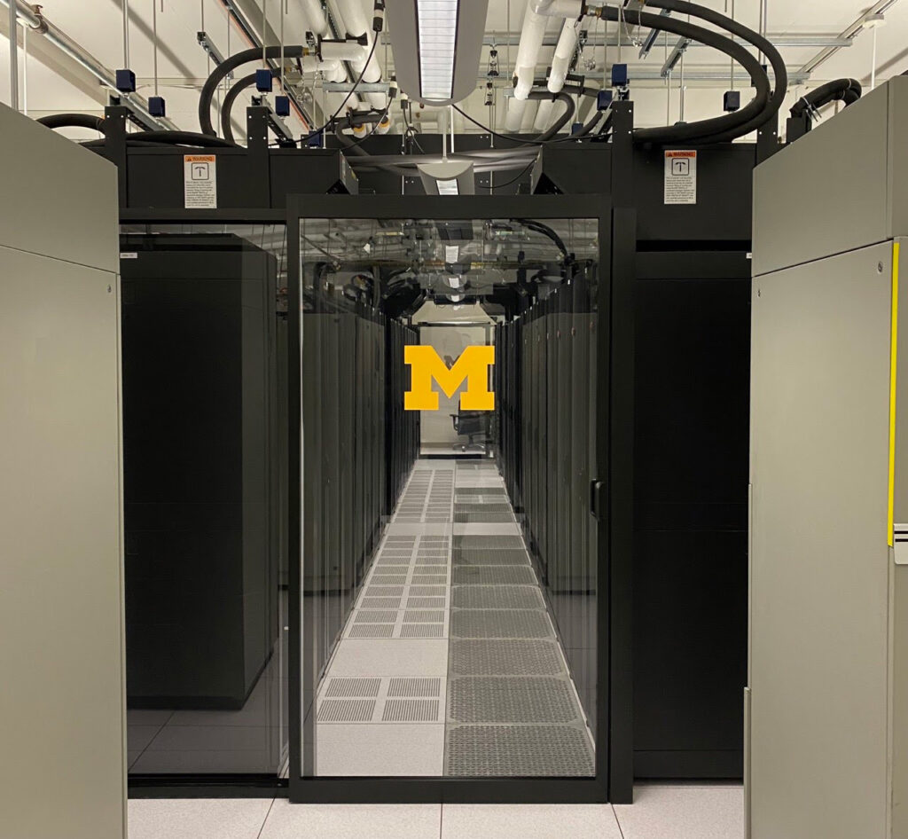 A glass door emblazoned with the U-M block M leads to a server room with black computer towers arranged in neat rows.