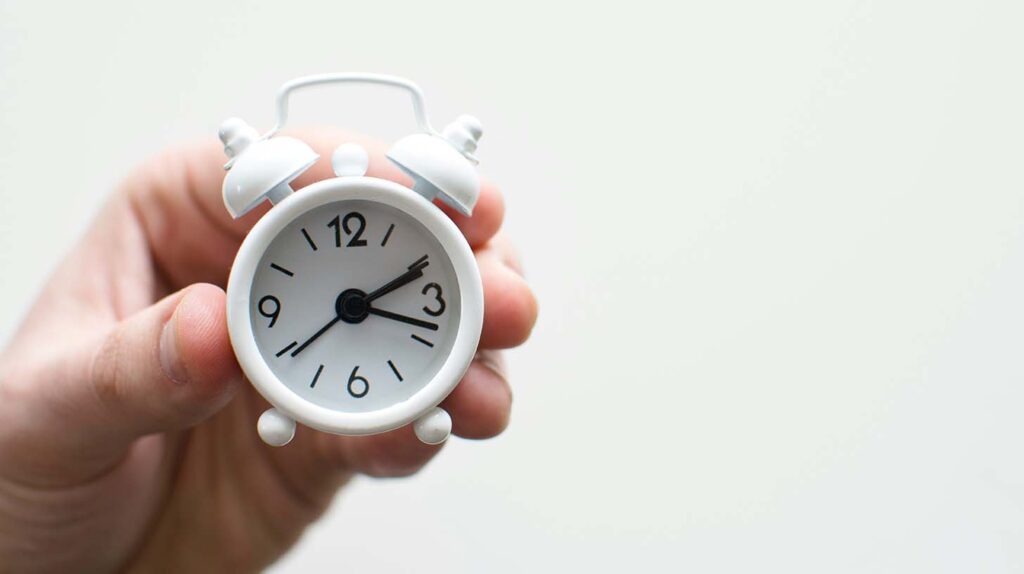 A hand holds a small analog alarm clock in front of a white background. Image credit: Lukas Blazek, Unsplash