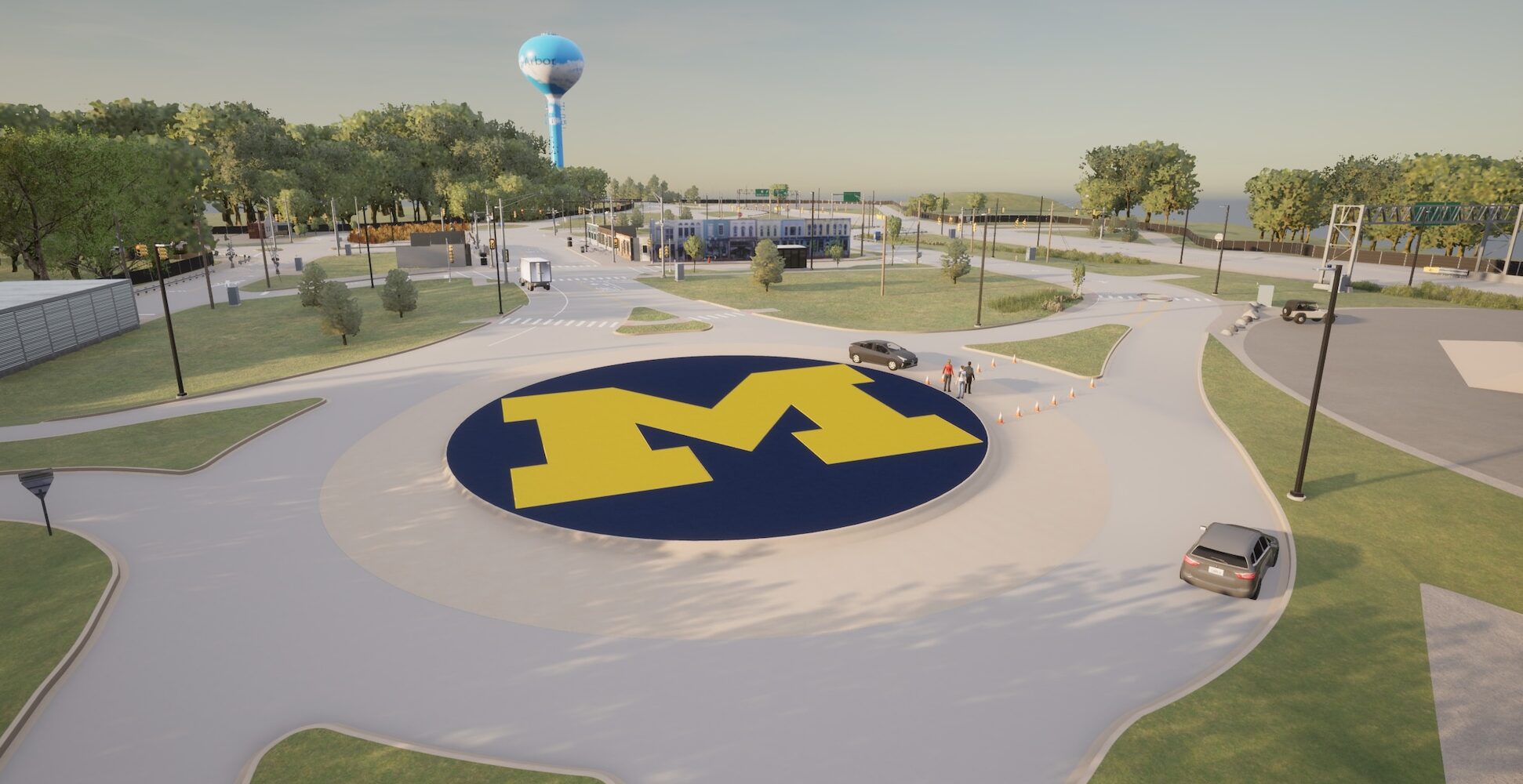 The simulation shows Mcity's roundabout with the block M in the center and green grass surrounding. The roundabout has multiple access roads and is partially blocked by a set of cones with some pedestrians nearby. More test track extends into the distance.