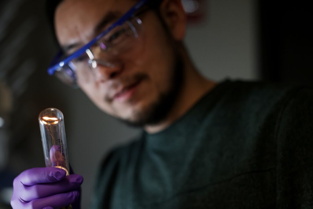 A filament glows golden at the end of a glass tube in Lu's gloved hand. He wears safety glasses.