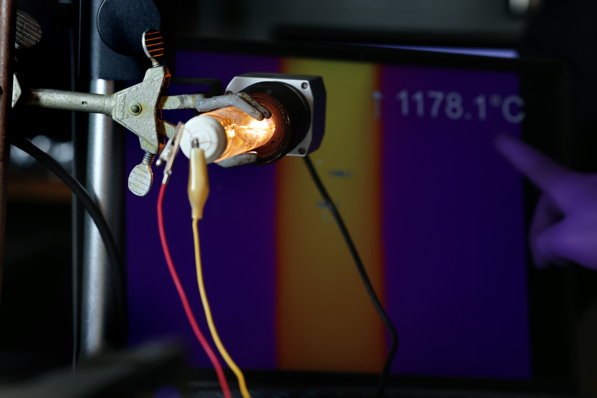Held in a metal clamp, the bulb glows golden with yellow and red wires attached to the rear. A gloved finger points to a temperature reading of 1178.1 degrees Celsius on the screen behind.Held in a metal clamp, the bulb glows golden with yellow and red wires attached to the rear. A gloved finger points to a temperature reading of 1178.1 degrees Celsius on the screen behind.