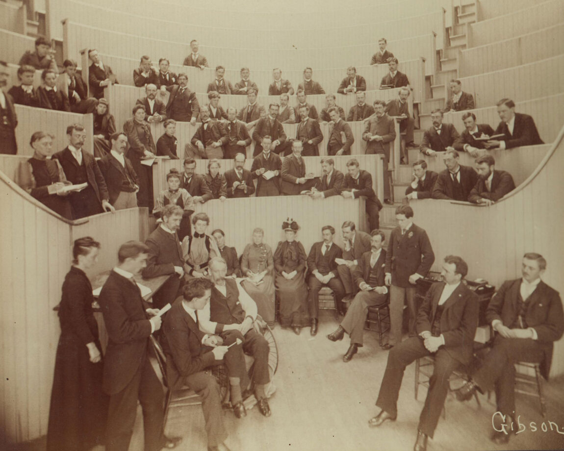 Photo showing early medical students and faculty with a patient in an amphitheater classroom. 