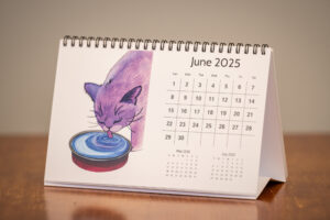 A photo of a calendar with a watercolor photo of a cat licking water out of a bowl