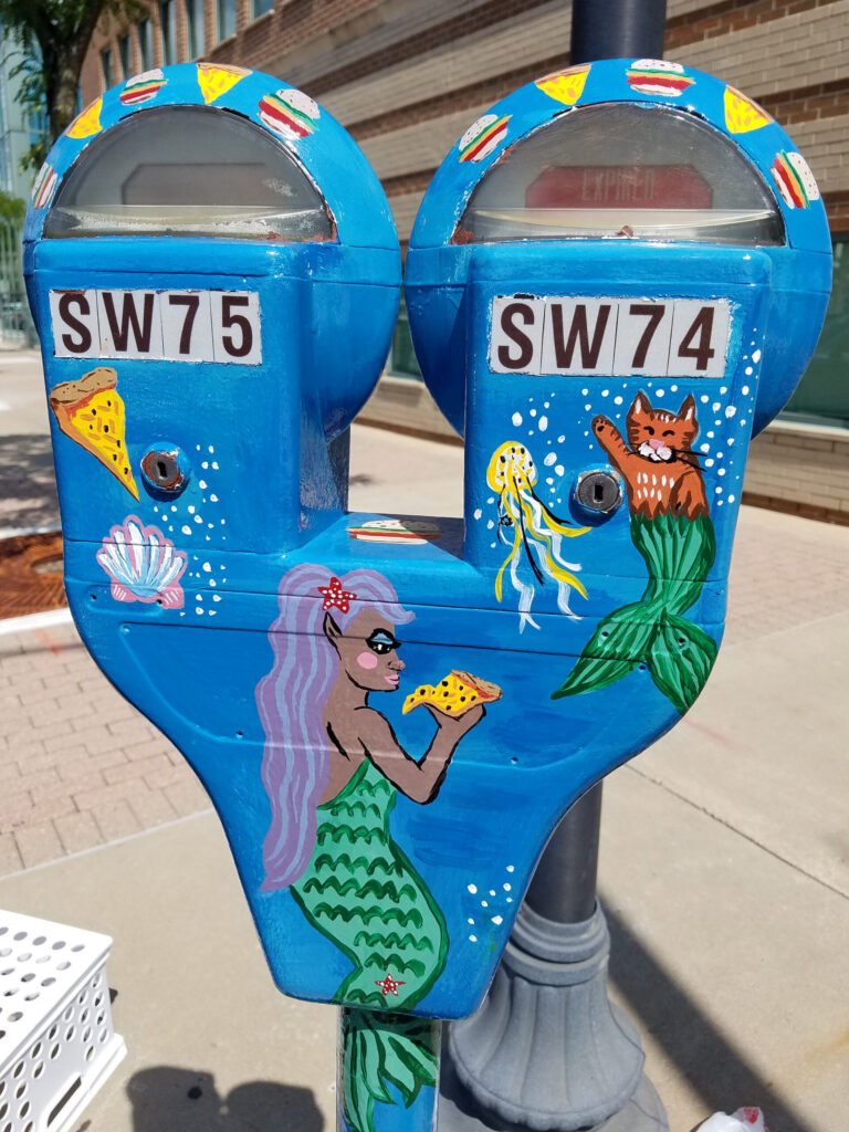 A parking meter painted with a scene depicting mermaids eating pizza