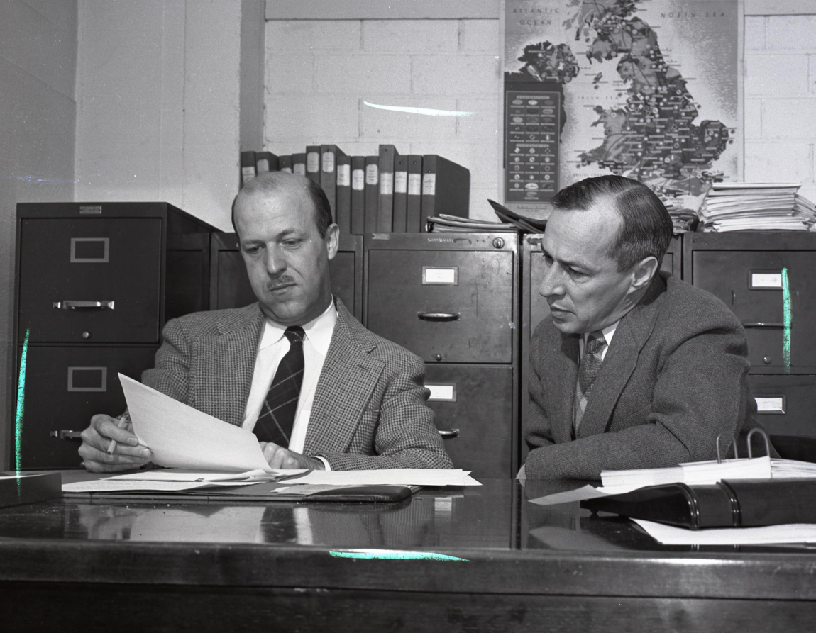 A photo of two men sitting at a desk looking at a piece of paper