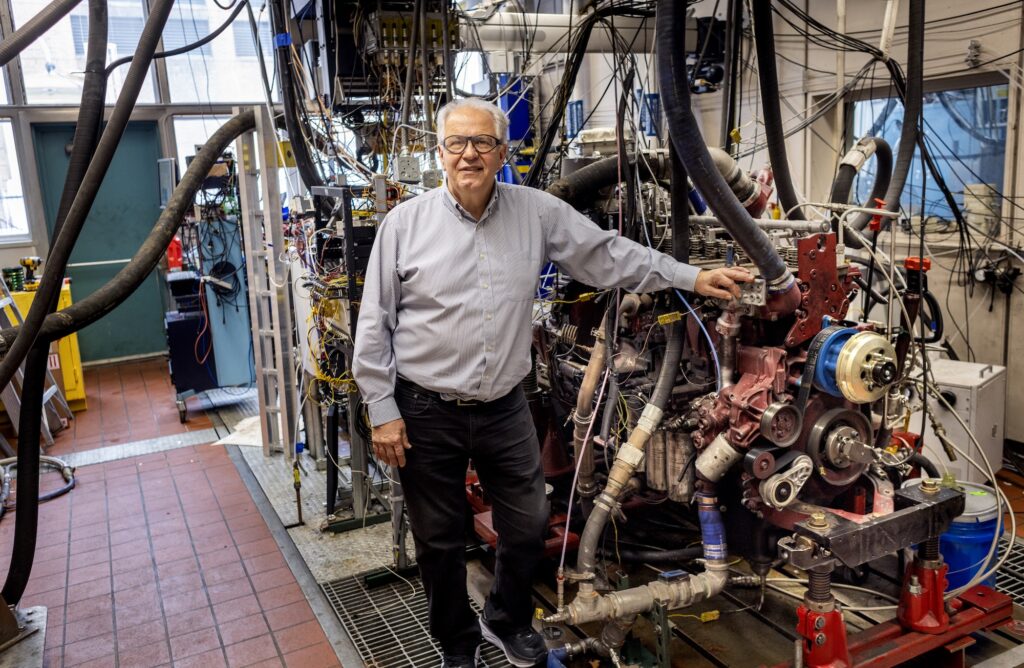 A man dressed in a button down shirt and dark pants stands in front of a massive engine inside a laboratory.
