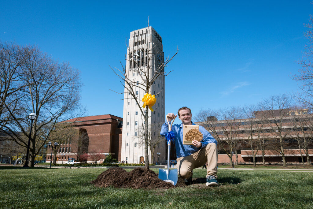 A man kneeling with a shovel and a plaque at the site where a young tree was planted