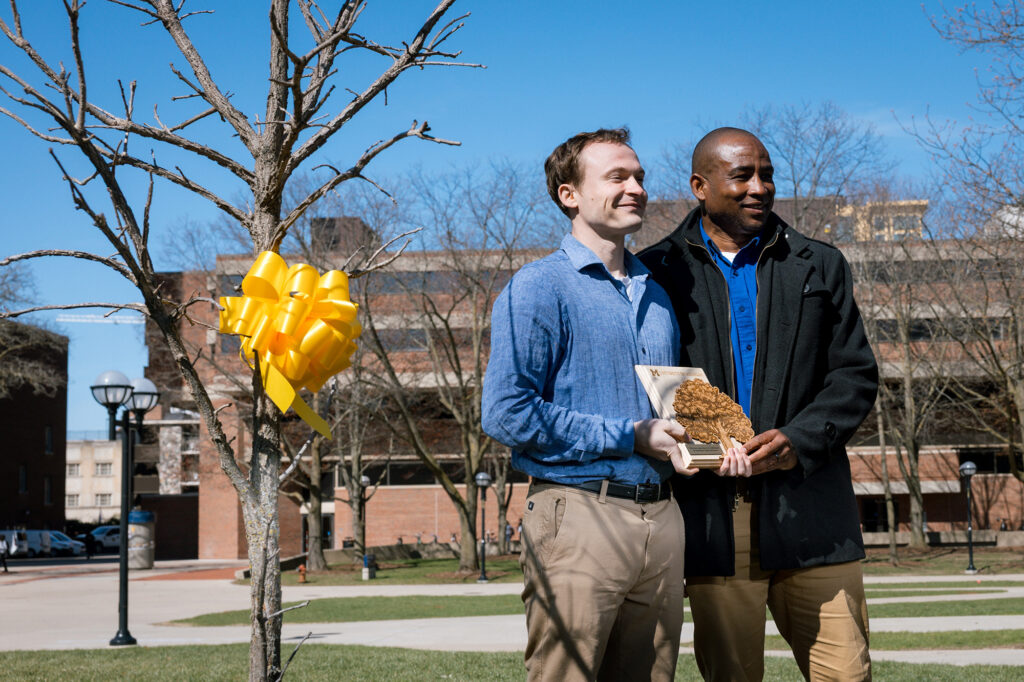 Two men smiling and holding a plaque with a small tree nearby